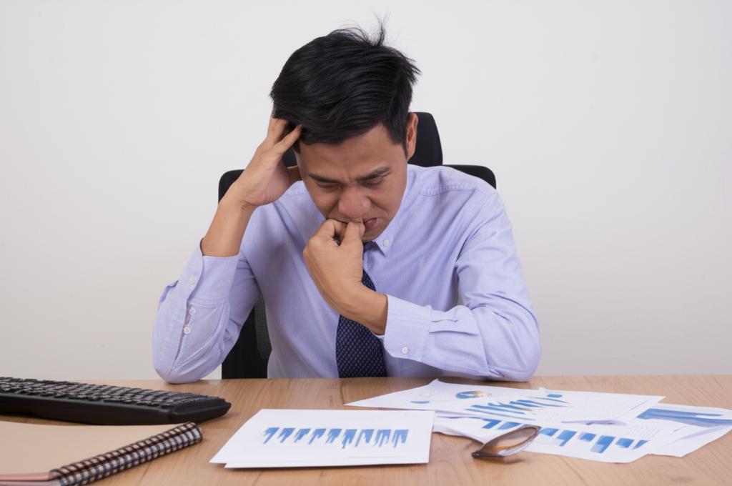 A stressed Thai businessman sitting at his desk, looking overwhelmed with paperwork, low-key lighting, realistic depiction.