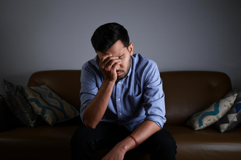 A man sitting alone on a sofa, looking stressed and thoughtful, with dim lighting, representing feelings of self-doubt and worry about performance issues.