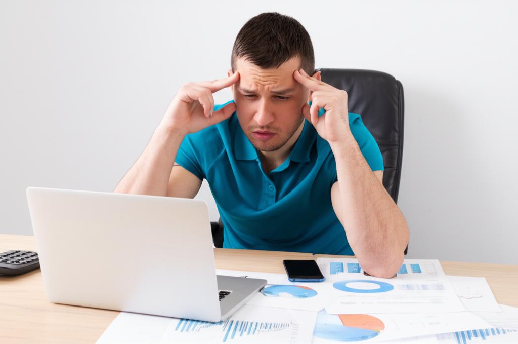 A young man sitting at a desk, looking tired and rubbing his temples, surrounded by work documents and a laptop, conveying stress and exhaustion.