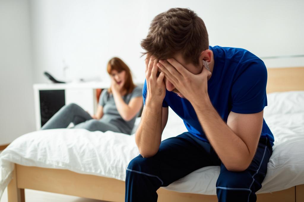 Man sitting on edge of bed looking tired and frustrated after exercise, partner looking concerned in background
